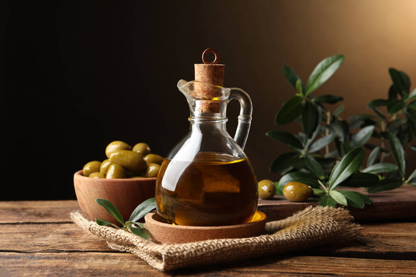 Glass jug of oil, ripe olives and green leaves on wooden table - 写真・画像