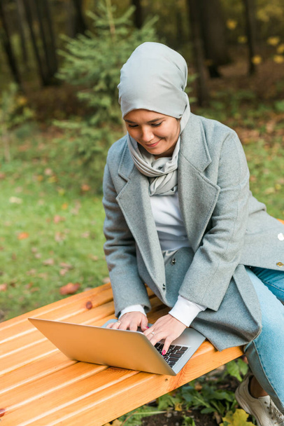 秋の公園でノートパソコンを使う若いアジア系ムスリム女性。オンライン教育、オンラインショッピング、ソーシャルネットワークの概念. - 写真・画像