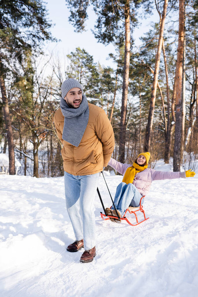Улыбающийся мужчина тянет сани с девушкой в зимнем парке  - Фото, изображение