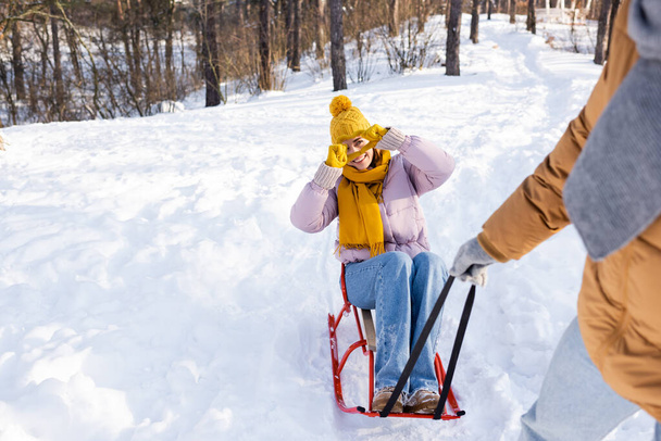 Smiling woman on sled showing love sign at blurred boyfriend in winter park  - Φωτογραφία, εικόνα