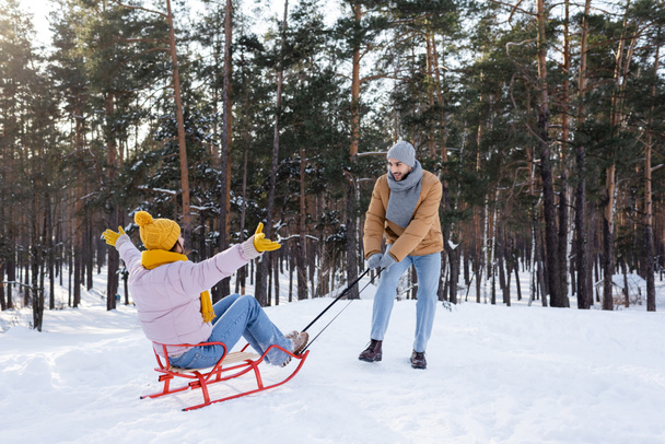glimlachende man trekken vriendin op slee op sneeuw in park  - Foto, afbeelding