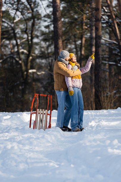 Side view of smiling man hugging girlfriend pointing away near sled in winter park  - Photo, Image
