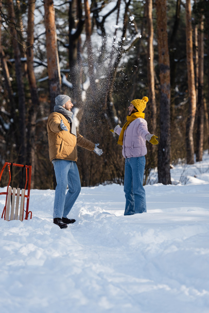 Vista lateral de pareja excitada en sombreros arrojando nieve cerca del trineo en el parque  - Foto, Imagen