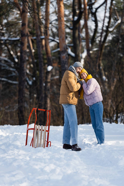Junges Paar lächelt beim Händchenhalten neben Schlitten im Winterpark  - Foto, Bild