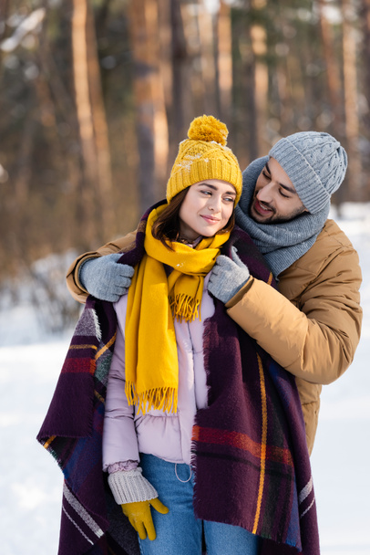 Smiling man wearing blanket on girlfriend in winter park  - Foto, Bild