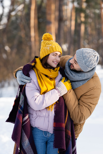 Smiling man hugging girlfriend with blanket in winter park  - Photo, Image