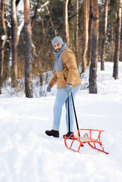 Улыбающийся мужчина в теплой одежде тянет сани в зимнем парке  - Фото, изображение