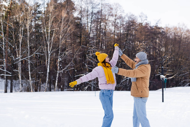 Side view of positive couple in warm hats holding hands and having fun in winter park  - 写真・画像
