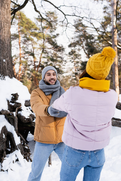 Wesoły mężczyzna trzymający za rękę dziewczynę na niewyraźnym pierwszym planie w zimowym parku  - Zdjęcie, obraz