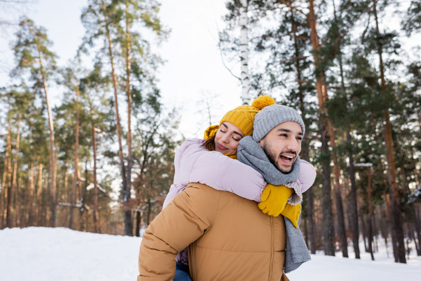 Woman with closed eyes embracing cheerful boyfriend in winter park  - Fotoğraf, Görsel