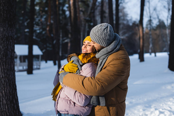 Muž v pleteném klobouku objímající hezké přítelkyně v zimním parku  - Fotografie, Obrázek