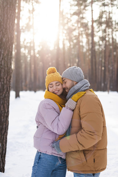 Sonriente hombre con los ojos cerrados de pie cerca de su novia en el parque de invierno  - Foto, Imagen