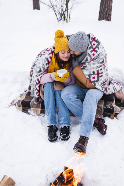 Young woman in winter outfit and blanket holding cup near boyfriend and bonfire on snow  - Photo, Image