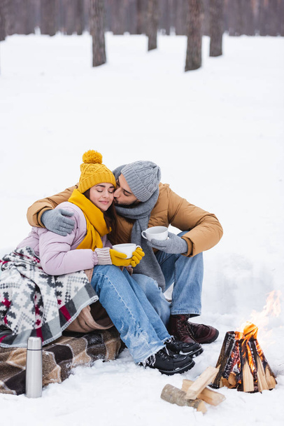 mężczyzna całuje dziewczyna z puchar i koc w pobliżu ognisko w zimie park  - Zdjęcie, obraz