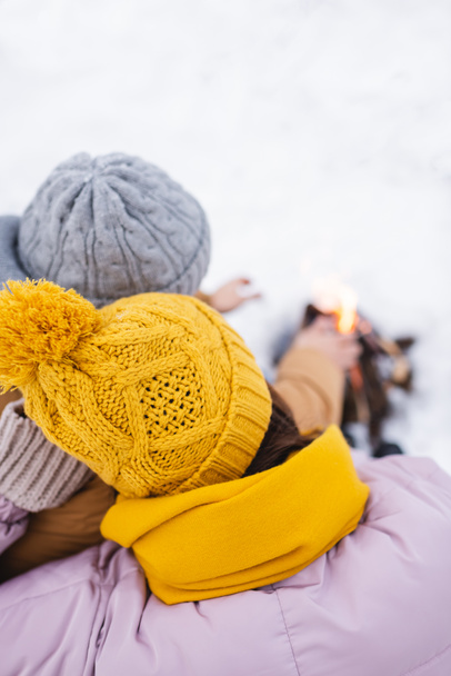 Γενική άποψη του ζευγαριού σε πλεκτά καπέλα κοντά σε θολή φωτιά στο χειμερινό πάρκο  - Φωτογραφία, εικόνα