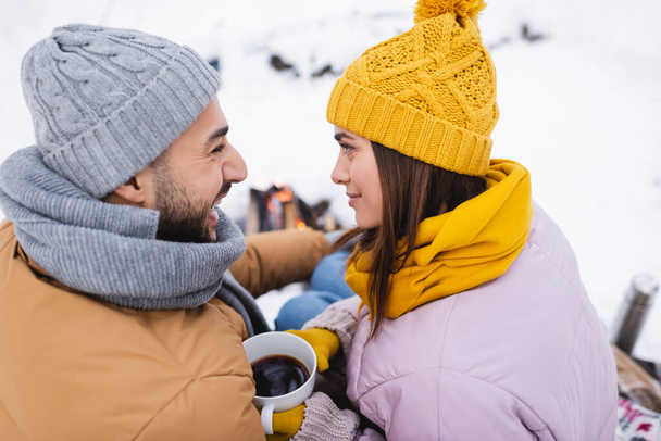 Side view of cheerful man looking at girlfriend with cup of coffee in winter park  - Foto, Imagen