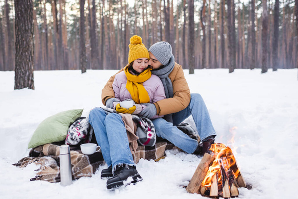 Man kissing girlfriend with cup near blankets and bonfire in winter park  - 写真・画像