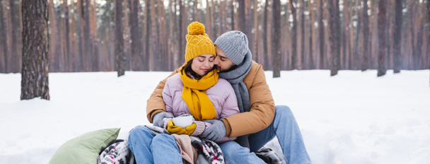 Młody mężczyzna całuje dziewczynę w stroju zimowym z filiżanką w zaśnieżonym parku, baner  - Zdjęcie, obraz