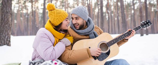 Pareja alegre en traje de invierno tocando la guitarra acústica en el parque, bandera  - Foto, Imagen