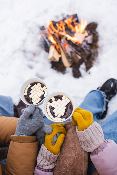 Top view of couple holding cups of cacao with marshmallows near blurred bonfire in winter park  - 写真・画像