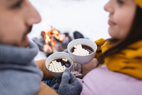 Cropped view of cups of cacao with marshmallows in hands of blurred couple in winter park  - Photo, Image