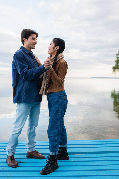 Vista lateral do casal interracial de mãos dadas no cais perto do lago  - Foto, Imagem