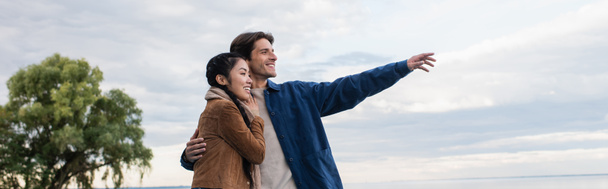 Fröhlicher Mann zeigt mit der Hand und umarmt asiatische Freundin mit See im Hintergrund, Banner  - Foto, Bild