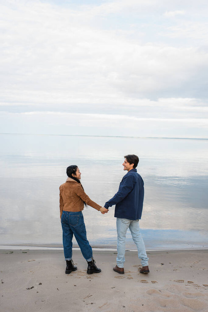 Side view of smiling interracial couple holding hands near sea during weekend  - Фото, изображение