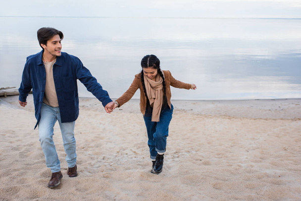 Smiling interracial couple holding hands while walking on beach  - Φωτογραφία, εικόνα