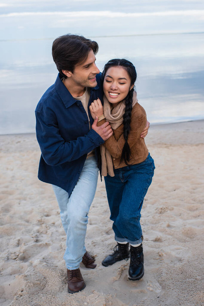 Man hugging cheerful asian girlfriend on beach  - Фото, зображення