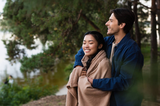 Smiling man hugging asian girlfriend in blanket outdoors  - Foto, imagen