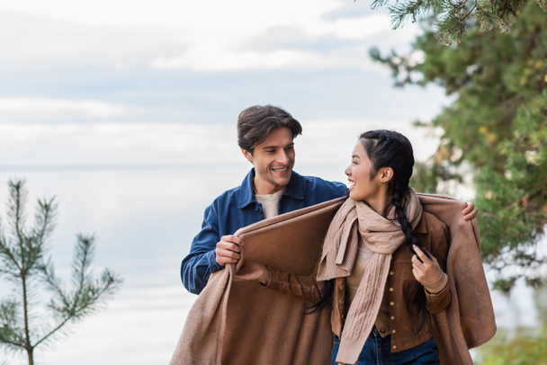 Man holding blanket near asian girlfriend with lake on background  - Fotó, kép