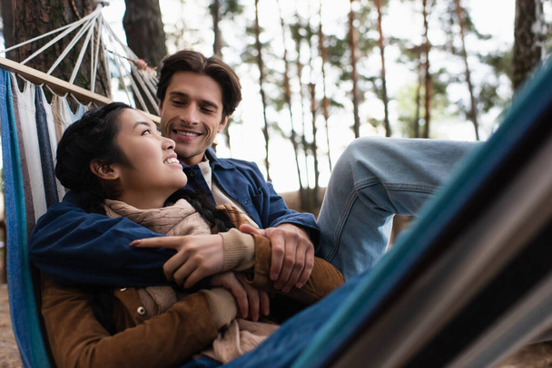 Cheerful man hugging asian girlfriend in blurred hammock outdoors  - Фото, изображение