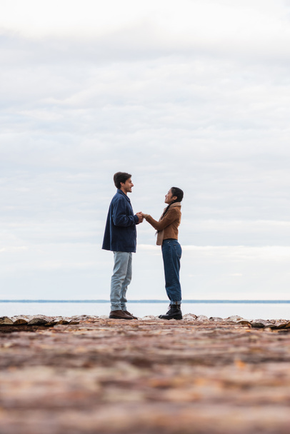 Side view of smiling interracial couple holding hands on pier near sea  - Photo, image