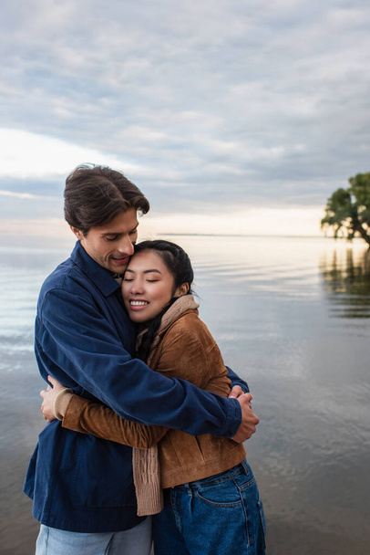 Smiling man hugging asian girlfriend with closed eyes near sea  - Zdjęcie, obraz