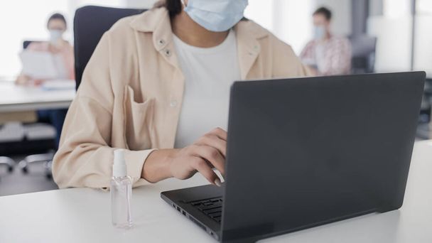 Young woman in protective mask working on laptop in business office, hand sanitizer on desk - Photo, image