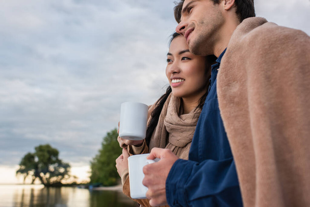Smiling multiethnic couple in blanket holding cups near blurred lake  - Foto, Imagen