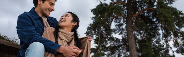 Low angle view of smiling man hugging asian girlfriend in blanket outdoors, banner  - Fotó, kép