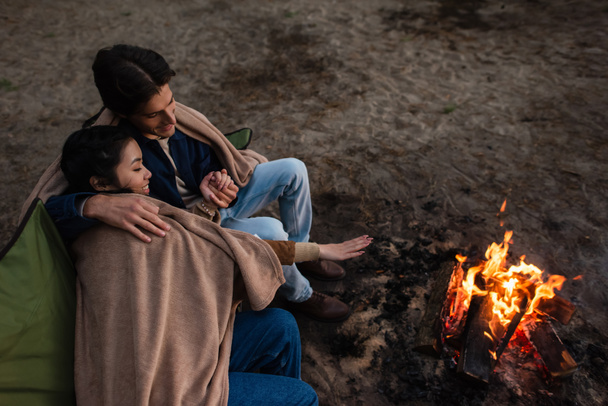 High angle view of smiling interracial couple in blankets looking at campfire  - Photo, Image