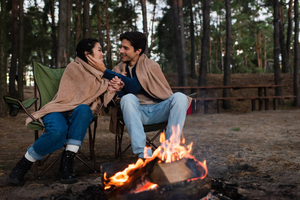 Smiling man touching face of asian girlfriend near blurred campfire outdoors  - 写真・画像