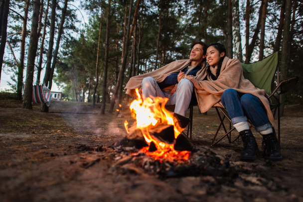 Young multiethnic couple in blankets sitting near blurred campfire  - Foto, immagini