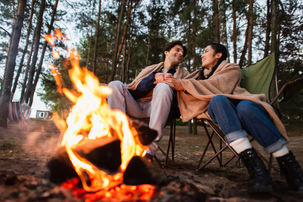 Multiethnic couple holding hands near blurred campfire during weekend  - Foto, Imagen