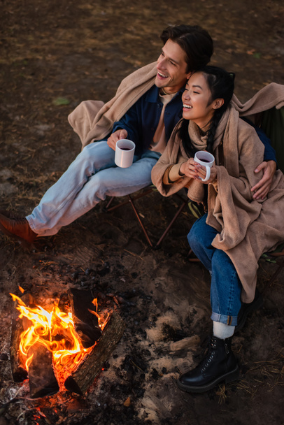 Smiling interracial couple with cups hugging near campfire during weekend  - 写真・画像