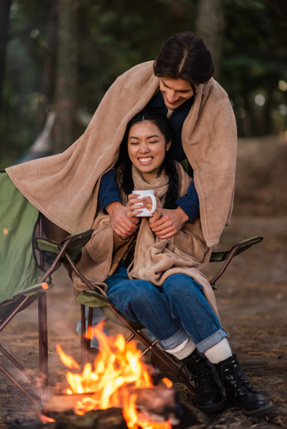 Bulanık kamp ateşinin yanında elinde fincanla Asyalı kız arkadaşının elini tutan bir adam.  - Fotoğraf, Görsel