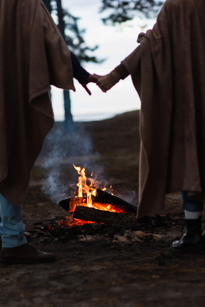 Vue recadrée du feu de camp près d'un couple flou se tenant la main le soir  - Photo, image