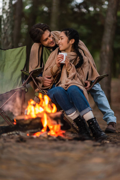 Smiling man in blanket standing near asian girlfriend with cup and blurred campfire on foreground  - 写真・画像
