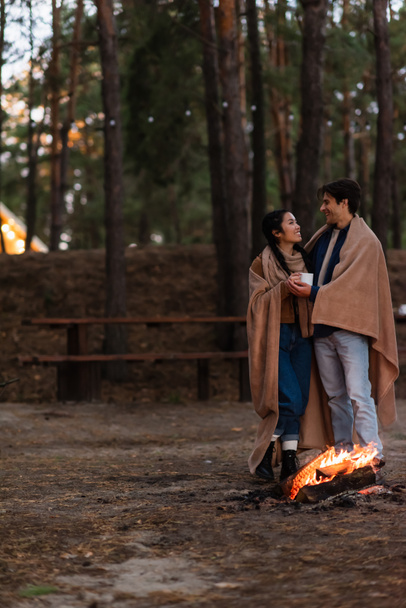 Couple interracial dans des couvertures avec tasse parler près de feu de camp dans la soirée - Photo, image