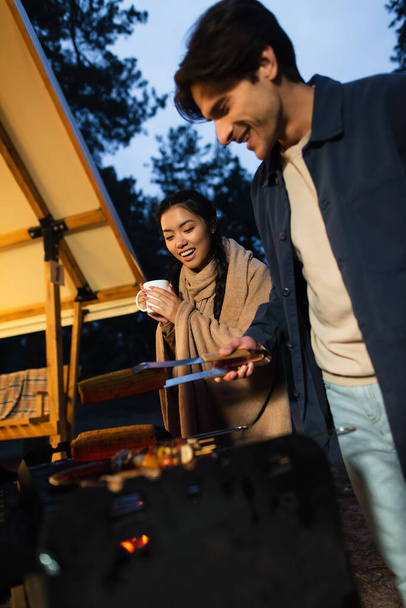Excited asian woman with cup standing near boyfriend grilling vegetables near glamping house outdoors  - Фото, зображення