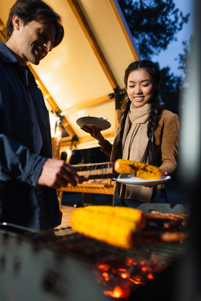 Asian woman holding plates near boyfriend and blurred grill outdoors  - Fotoğraf, Görsel