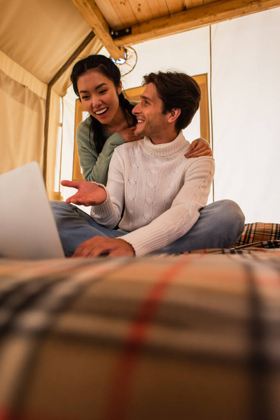 Smiling man pointing at laptop near asian girlfriend in glamping house  - Foto, immagini